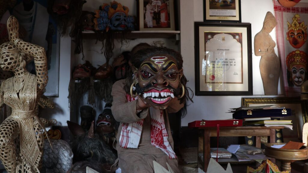 Mask maker holding large painted mask inside workshop surrounded by crafts.