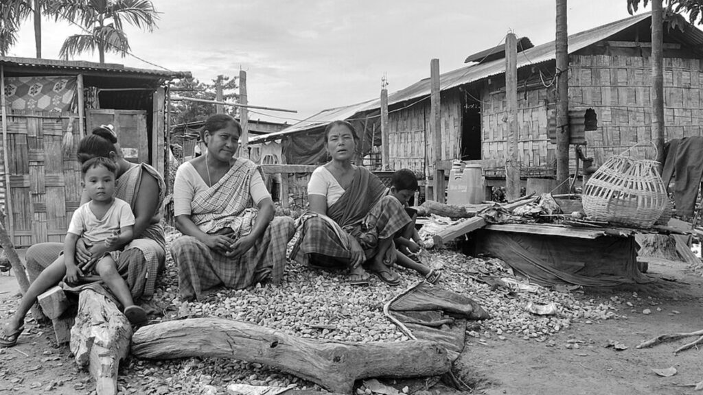 Mising women and children sitting outside bamboo houses in village.