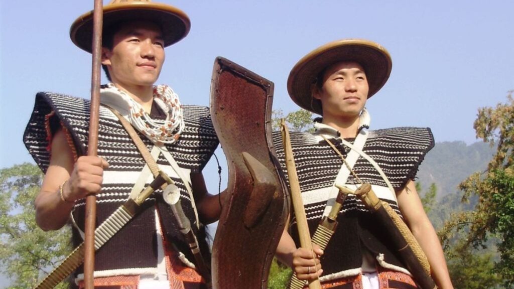 Two Idu Mishmi men in traditional black-and-white attire holding weapons and shields.