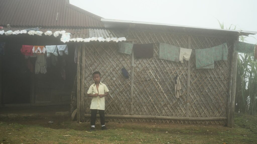 Young boy standing outside bamboo house on misty morning.