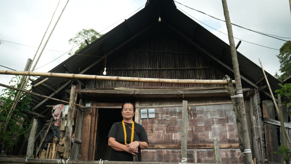 Woman stands proudly before bamboo house.