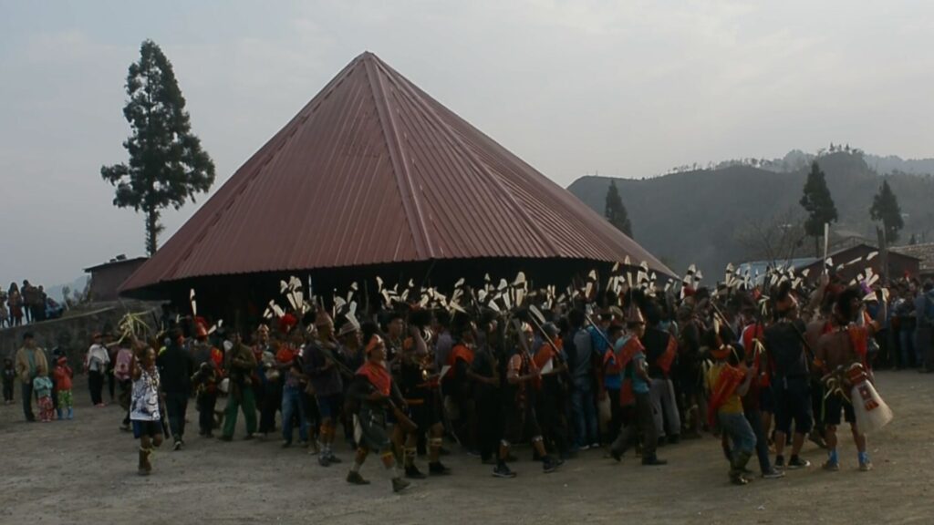 Large gathering of Konyak warriors dancing around morung house.