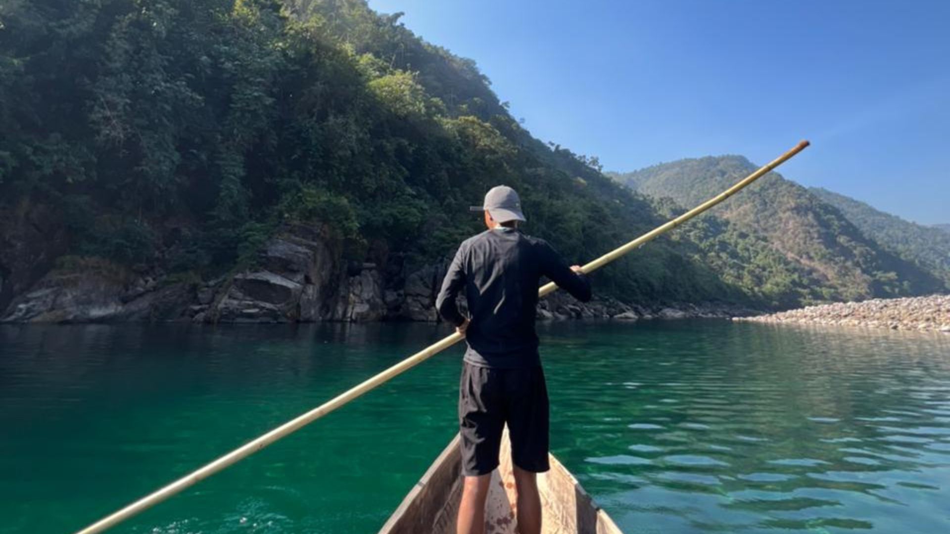 Boatman on a wooden canoe poling across emerald river with forested hills under blue sky in Disong, Meghalaya.