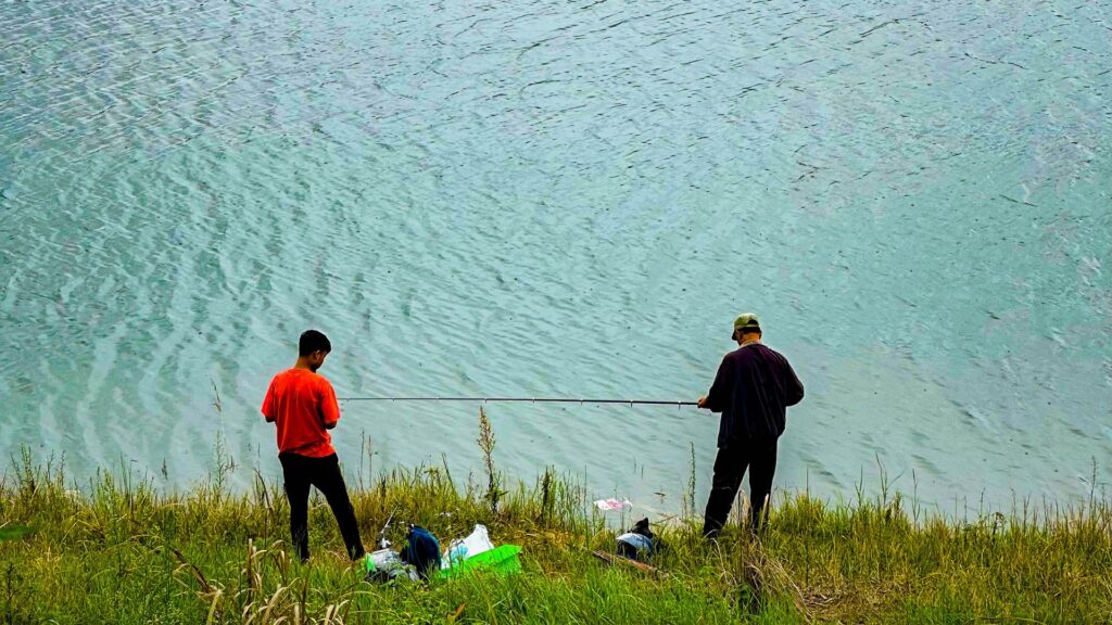 Two anglers fishing from a grassy bank above turquoise water